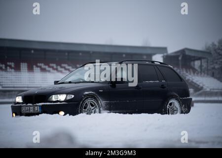 12-12-2022 Riga, Lettland ein schwarzes Auto parkt im Schnee in der Nähe eines Stadiongeländes mit einem Gebäude im Hintergrund und einem schneebedeckten Feld. . Stockfoto