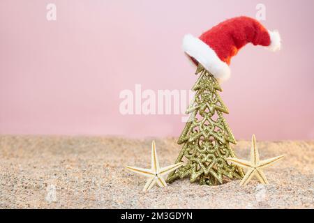 Goldener Weihnachtsbaum mit Weihnachtsmannmütze und Seesternen auf Sand. Weihnachten, Neujahr. Speicherplatz kopieren Stockfoto