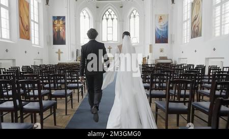 Wunderschöne Frischvermählte, die den Gang entlang gehen. Aktion. Rückansicht von zwei frisch Vermählten in der Kirche. Frisch Vermählte gehen bei der Hochzeitspräferenz vor den Altar. Stockfoto