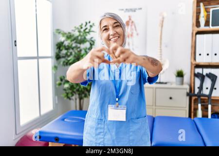 Grauhaarige Frau im mittleren Alter, die Physiotherapeut-Uniform in der medizinischen Klinik trägt, in Liebe lächelt und Herzsymbol mit Händen formt. Romantisches Konzept. Stockfoto