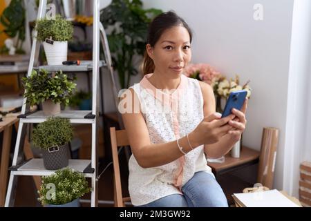 Junge asiatische Blumenfrau, die selbstbewusst mit ihrem Smartphone im Blumenladen lächelt Stockfoto