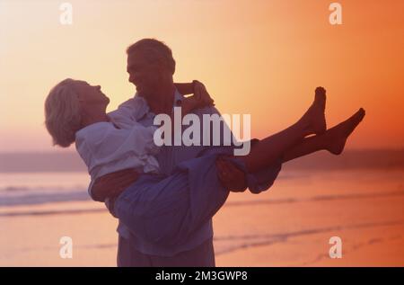 Ein älteres Paar, das vor dem Sonnenuntergang am Strand herumschnüffelt. Frau in Männerarmen. Stockfoto