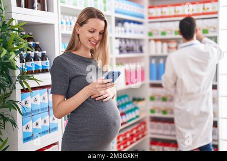 Mann und Frau, Apotheker und schwangere Klientin, die in der Apotheke ein Smartphone benutzen Stockfoto