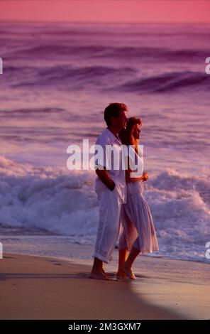 Ein junges Paar in Weiß auf einem romantischen Spaziergang am Strand bei Sonnenuntergang Stockfoto