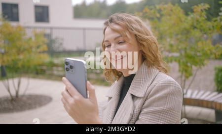 Schöne Frau kommuniziert per Video auf der Straße. Aktion. Eine stilvolle Frau spricht und lächelt über einen Videolink. Glückliche Frau, die mit Video telefoniert. Stockfoto