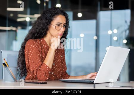Ernsthaft denkende hispanische Geschäftsfrau, die in einem modernen Büro arbeitet, weibliche Programmiererin, die auf einem Laptop neue Software testet. Stockfoto