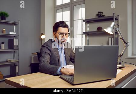 Fokussierter, selbstbewusster Geschäftsmann, der in einem modernen Büro mit einem Notebook arbeitet Stockfoto