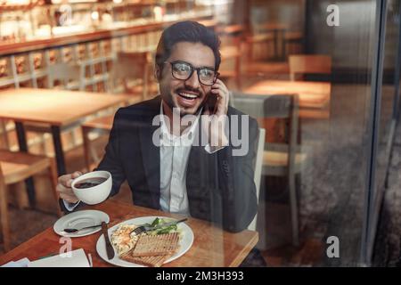 Ein lächelnder indischer Geschäftsmann spricht während der Mittagszeit in einem gemütlichen Café mit einem Kunden Stockfoto