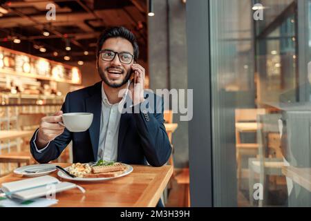 Ein gutaussehender indischer Geschäftsmann mit Brille spricht während der Mittagspause in einem gemütlichen Café mit dem Kunden Stockfoto
