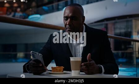 Ethnischer afroamerikanischer Geschäftsmann mittleren Alters im Café Tisch reifer Mann CEO mit Handy Geschäft plaudern entspannend essen Kuchen essen Kuchengetränk Stockfoto