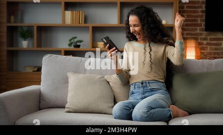 Glückliche Frau, die das Telefon für Wetten im Internet benutzt, erhalten Nachricht mit guten Nachrichten Angebot zu Hause auf der Couch zu sitzen, gewinnen das Handy-Gewinnspiel online Stockfoto