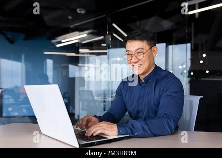 Ein fröhlicher und lächelnder asiatischer Mann, der im Büro arbeitet, ein Geschäftsmann mit Hemd und Brille, der ein Notebook benutzt, ein Entwickler, ein Programmierer, der Code-Programmiersoftware eingibt. Stockfoto