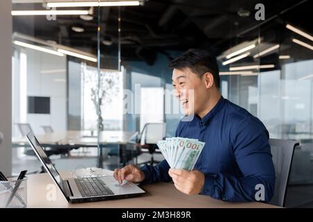 Asiatischer Geschäftsmann, Investor, der mit Erfolg und finanziellen Erfolg zufrieden ist, Mann, der in einem modernen Büro arbeitet und ein Notebook am Arbeitsplatz benutzt, Geld in bar in US-Dollar hält. Stockfoto