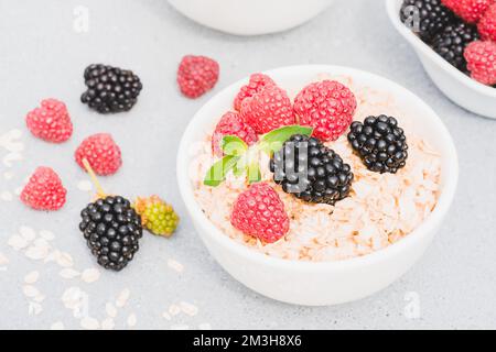 Haferbrei garniert mit Beeren und Minzblättern in einer weißen Schüssel auf dem Tisch. Morgencreme für Superfood. Ideen für ein gesundes Frühstück Stockfoto