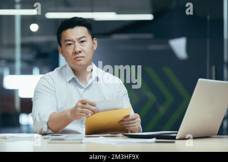 Ein seriöser und angepekter gutaussehender Mann, ein asiatischer Geschäftsmann, sitzt im Büro am Tisch und hält einen Umschlag mit einem Brief in der Hand. Schlechte Nachrichten, finanzielle Probleme, Rechnungen. Stockfoto