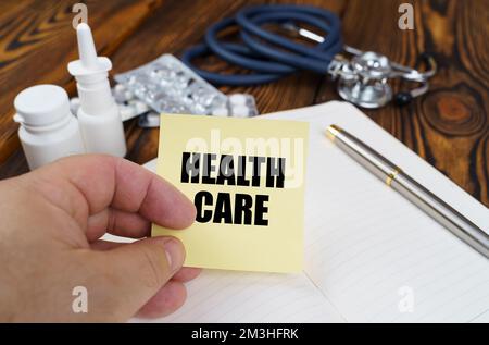 Medizinisches Konzept. Auf dem Tisch liegt ein Notizbuch, Medikamente und ein Stethoskop, in den Händen eines Aufklebers mit der Aufschrift - HEALTH CARE Stockfoto