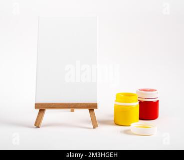 Vertikale Leinwand, leeres Papier auf Holzständer, Gouachenfarben in Gläsern, rot und gelb. Hochwertiges Foto Stockfoto