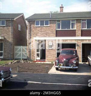 cira Ende 1960er, historisch, ein kastanienbrauner Humber Hawk Wagen aus der Nachkriegszeit, der in der Einfahrt eines neuen, halbseparaten Hauses geparkt wurde, mit einer internen Garage, erbaut im modernen Stil der Epoche, wie er zu dieser Zeit auf vielen Wohnhäusern in Großbritannien zu sehen war. Der Humber Hawk wurde zwischen 1945 und 1967 von Humber Limited in Britiain gebaut und bietet verschiedene Stile und Varianten. Das Auto, das hier zu sehen ist, ist sehr wahrscheinlich der Humber Hawk Mark VI, der 1954 eingeführt wurde, eine aktualisierte Version des Nachkriegs-1948 Hawk, ein komplett neu gestaltetes Auto als der erste und ursprüngliche 1945 Hawk. Stockfoto