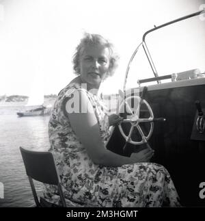 1960er, historisch, eine Dame sitzt am Steuer, T das Bootsrad eines Motorstarts, das in einem Hafen in England, Großbritannien, festgemacht ist. Stockfoto