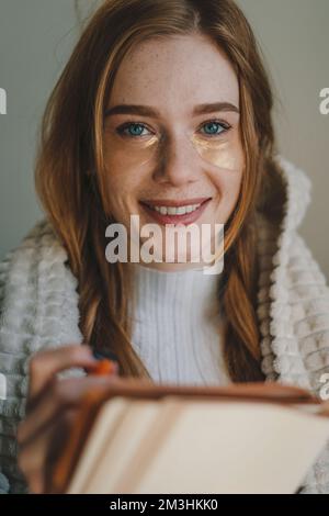 Porträt einer Frau, die in die Kamera lächelt, mit Sommersprossen und rotem Haar, mit blauen Augen, mit einem Notizbuch in der Hand, in dem sie ihre Gedanken schreibt Stockfoto