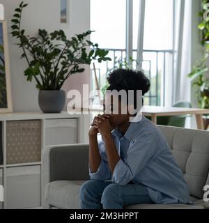 Unterdrückte afroamerikanische Frau mit traurigem Gesicht sitzt auf dem Sofa und denkt an die Zukunft. Lebensgefährliche Probleme. Stockfoto