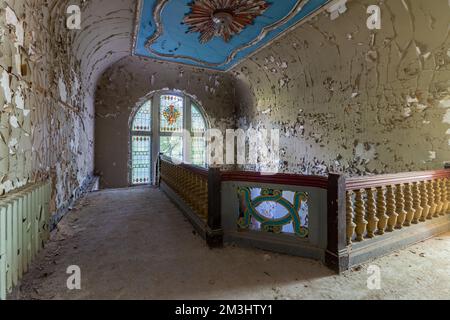 Ein mystischer Blick auf eine Holztreppe in einem alten Herrenhaus mit abblätternder Farbe an der Wand, einer wunderschönen blauen Decke und einem kunstvoll verzierten Fenster Stockfoto