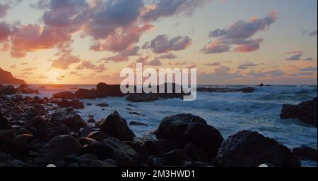 Meereswellen, die nördlich der Insel Teneriffa gegen Felsenklippen stürzen. Wellen brechen bei Sonnenuntergang auf schwarzen Steinen. Schäumende blaue Ozeanwellen. Stockfoto