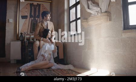Elegantes Paar am Fenster im alten Haus. Aktion. Ein paar Liebende umarmen sich am Fenster. Ein wunderschönes Paar, das sich umarmt und aus dem Fenster schaut. Stockfoto