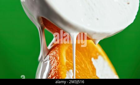 Nahaufnahme des Gießens von weißer Glasur auf zwei Hälften einer geschnittenen Orange. Clip. Saftige Früchte mit süßer Glasur, isoliert auf grünem Hintergrund Stockfoto