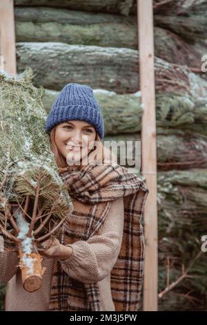 Ein hübsches Mädchen mit blauer Mütze und Schal trägt einen Weihnachtsbaum auf der Schulter. Ein Mädchen, das zu Weihnachten gekleidet ist, bringt eine frisch geschnittene Tanne mit nach Hause Stockfoto