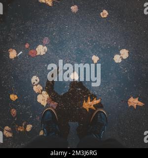 Eine Nahaufnahme von Personenfüßen auf nassem Asphalt mit Reflexion und Herbstblättern Stockfoto