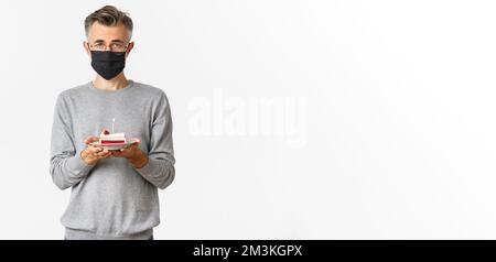 Konzept von Coronavirus, Quarantäne und Urlaub. Bild von traurigen Mann mittleren Alters in schwarzer medizinischer Maske und Brille, hält Geburtstagskuchen mit brennenden Kerze Stockfoto