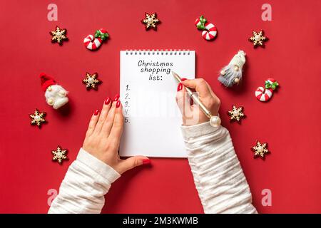 Weibliche Hand hält Stift und schreibt Weihnachtseinkaufsliste, Geschenkideen auf weißem Notizblock auf rotem Hintergrund Top View flach liegend Weihnachtskarte Frau macht C Stockfoto