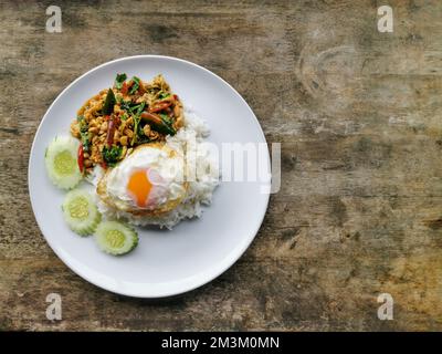 kra pao Gai auffüllen oder gebratenes Hähnchen mit thailändischem Basilikum, Spiegelei und weißem Reis auf weißem Teller mit Holzhintergrund umrühren. Street Food in Thailand. Stockfoto