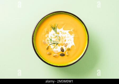 Dicke Butternusskürbis-Bisque in grüner Schüssel garniert mit schwerer Sahne, Rosmarin und Samen auf hellgrünem Hintergrund. Schale mit Kürbiscreme-Suppe. Vi Stockfoto