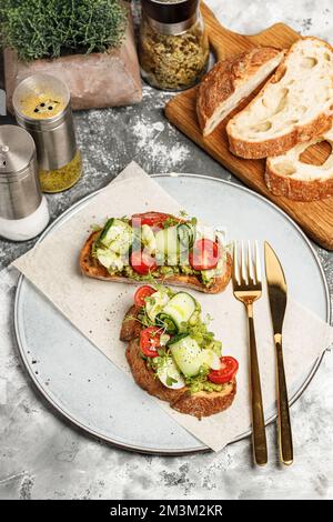 Sandwiches mit Guacamole-Sauce und frischem Gemüse auf einem runden Teller. Toasts mit Avocado, Gurke, Kirschtomaten und Kräutern. Vegetarisches Frühstück Stockfoto