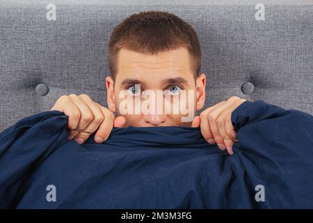 Direkt in den Rahmen schauen. Ein hübscher junger Mann, der sich hinter einer Decke versteckt, schaut zur Seite. Blickrichtung in verschiedene Richtungen. Stockfoto