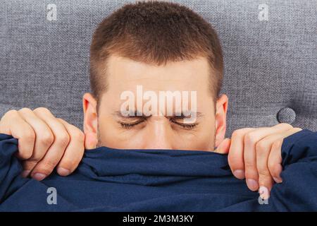 Mit geschlossenen Augen. Ein hübscher junger Mann, der sich hinter einer Decke versteckt, schaut zur Seite. Blickrichtung in verschiedene Richtungen. Stockfoto