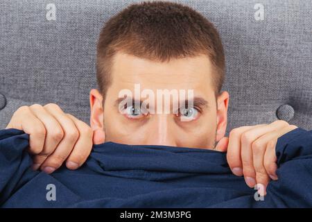 Direkt in den Rahmen schauen. Ein hübscher junger Mann, der sich hinter einer Decke versteckt, schaut zur Seite. Blickrichtung in verschiedene Richtungen. Stockfoto