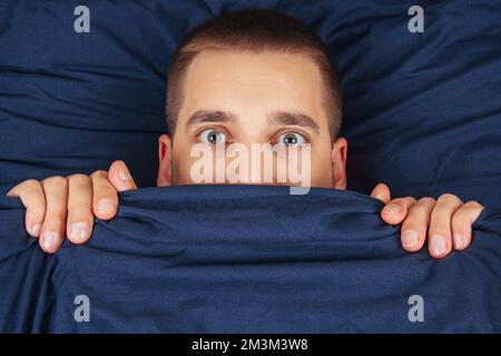 Direkt in den Rahmen schauen. Ein hübscher junger Mann, der sich hinter einer Decke versteckt, schaut zur Seite. Blickrichtung in verschiedene Richtungen. Stockfoto