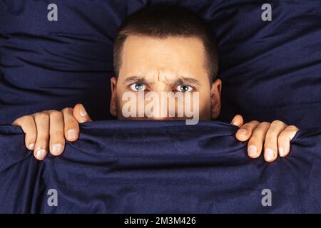 Direkt in den Rahmen schauen. Ein hübscher junger Mann, der sich hinter einer Decke versteckt, schaut zur Seite. Blickrichtung in verschiedene Richtungen. Stockfoto