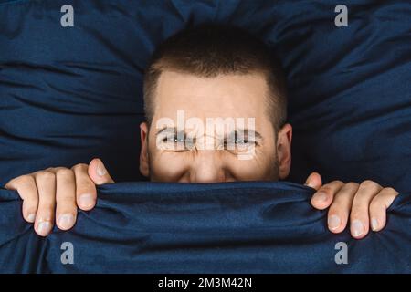 Direkt in den Rahmen schauen. Ein hübscher junger Mann, der sich hinter einer Decke versteckt, schaut zur Seite. Blickrichtung in verschiedene Richtungen. Stockfoto