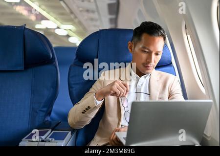 Professioneller und fokussierter asiatischer Geschäftsmann in einem formellen Business-Anzug hält seine Brille und benutzt während des Fluges einen Laptop. Stockfoto