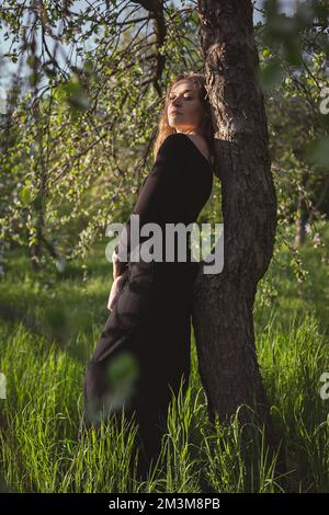 Junge Frau in schwarzem Kleid lehnt sich gegen die malerische Fotografie von Bäumen Stockfoto