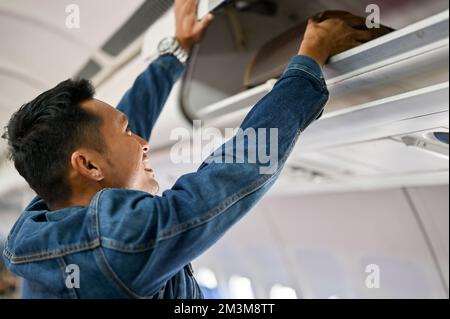 Der attraktive männliche asiatische Passagier nimmt sein Handgepäck nach der Landung am Zielort aus dem Gepäckfach. Transportkonzept Stockfoto