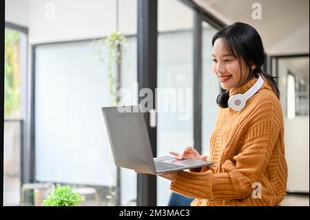 Hübsche und charmante junge Asiatin steht im Raum und benutzt ihren tragbaren Laptop. Stockfoto