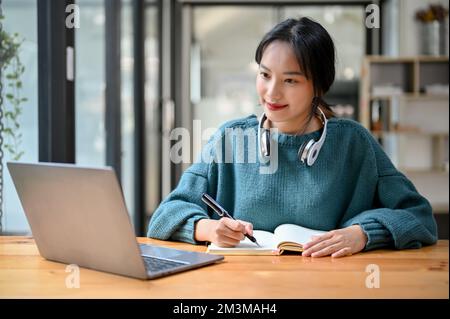 Hübsche und charmante junge asiatische Studentin, die sich auf ihre Hausaufgaben oder ihr Schulprojekt konzentriert, auf den Laptop-Bildschirm schaut und sich Notizen auf ihrer Schule macht Stockfoto