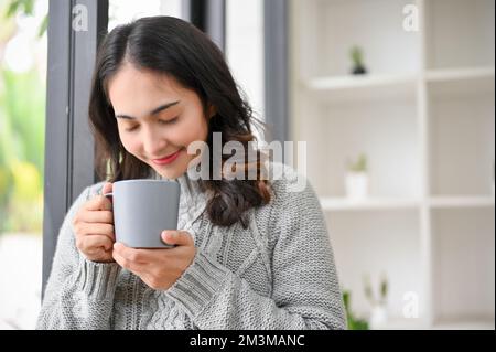 Friedliche und ruhige Asiatin der Jahrtausendwende in einem gemütlichen Pullover, die ihren Morgenkaffee genießt. Aroma-, Kaffee- oder Teepause Stockfoto