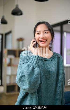 Porträt, schöne junge Asiatin in einem gemütlichen Pullover, spricht gern mit ihrer Freundin am Telefon. Stockfoto