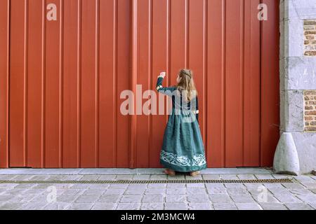 Ein kleines Mädchen in mittelalterlichen Kleidern, das an das große rote Tor des alten Schlosses klopft Stockfoto